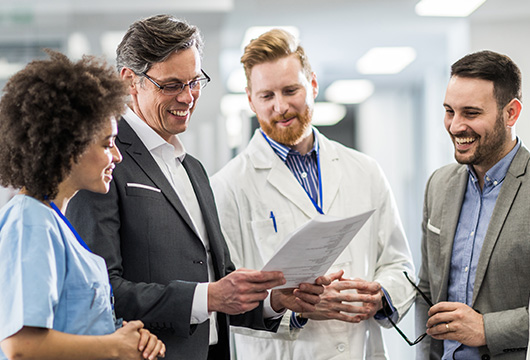 An image of two males in suits talking with a male doctor in a white lab coat and a female nurse in scrubs