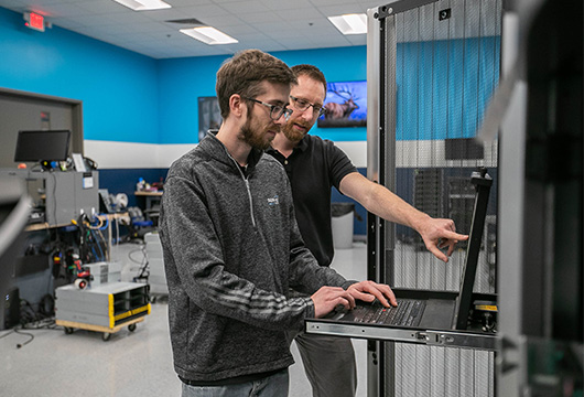 An image of to male engineers in a data center one is standing at a laptop and the other is behind him pointing to the screen